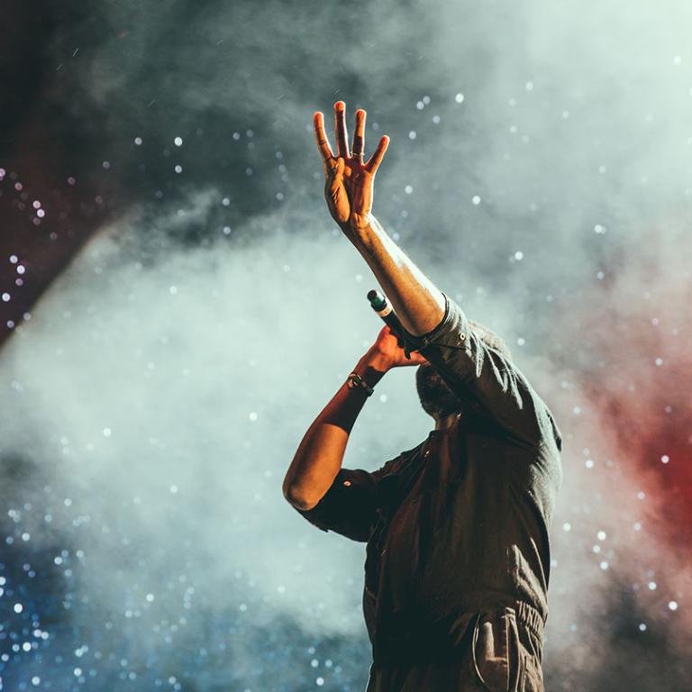 En artist sjunger i mikrofon och lyfter en hand mot skyn, mot en bakgrund fylld med scenrök