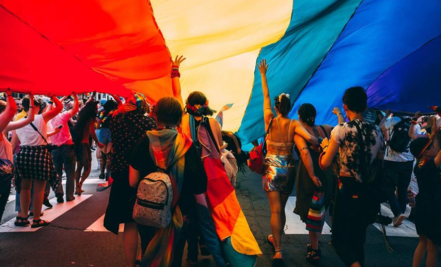 Bild på människor som står under en regnbågsflagga. Fotograf: Mercedes Mehling