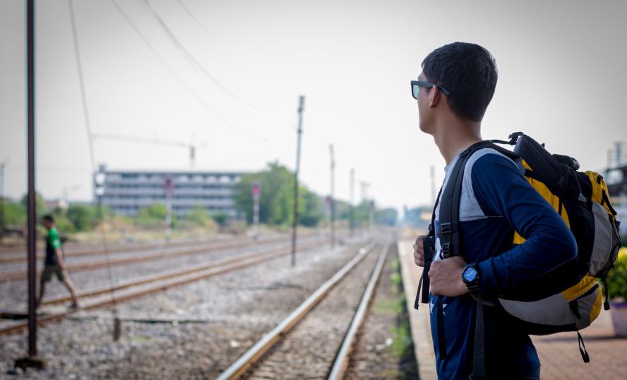 ung person står med ryggsäck på en tågperrong och blickar bort från kameran mot tågspåret.