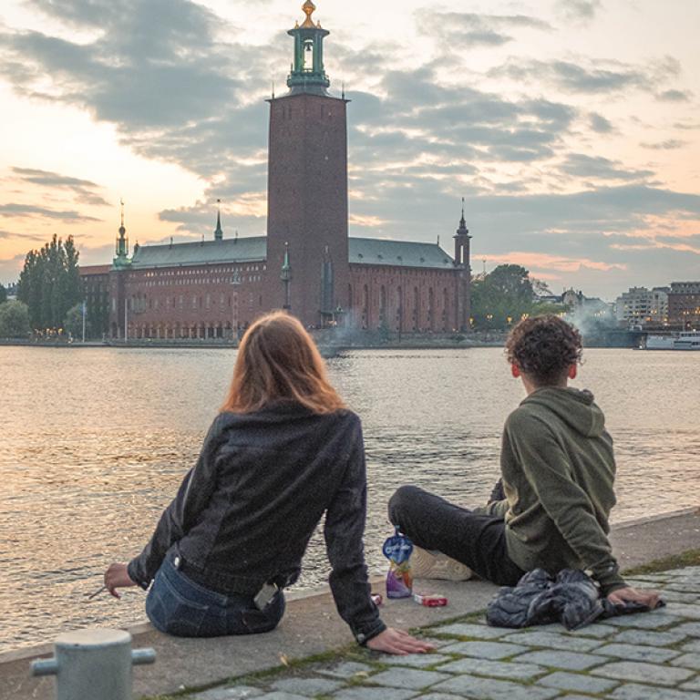 Bild på två unga personer som sitter vid vattnet och blickar mot Sveriges stadshus. Fotograf: Nikola Johnny Mirkovic