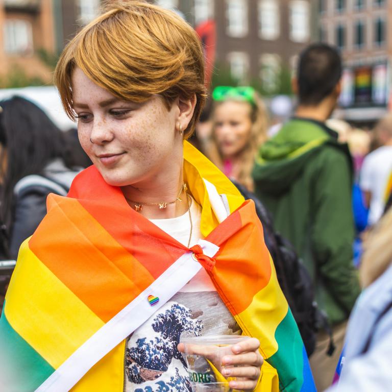 Ung person i en folksamling med prideflagga över axlarna. Foto: Jose Maria Sava/Unsplash